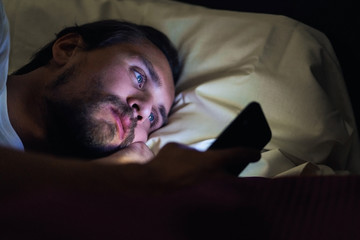 Young handsome and tired man with a beard cannot sleep and is watching something on his phone at...