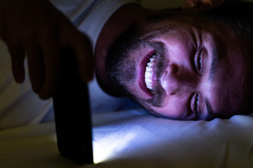 Bearded young handsome and tired man cannot sleep and is watching something on his phone at night. Blue light from the phone is all over his face.