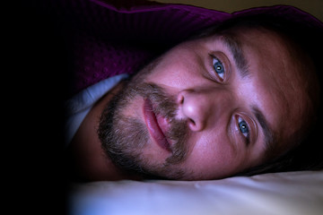 Bearded young man is lying in bed under his blanket. He cannot sleep and is watching something on...