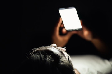 Young man is lying in his bed at night while watching something on his mobile phone.