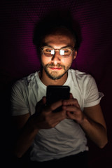 Bearded young man is lying in his bed at night while watching something on his mobile phone. Phone's reflection is visible in man's glasses.