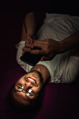 Bearded young man is lying in his bed at night while watching something on his mobile phone. Phone's reflection is visible in man's glasses.