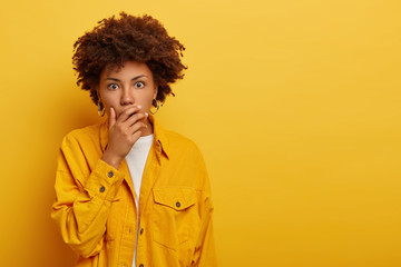 Fototapeta na wymiar Studio shot of scared curly haired female covers mouth with fright, scared by terrifying thing, dressed in fashionable yellow jacket, cannot believe eyes, stands indoor, blank copy space for advert