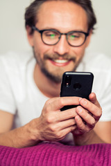 Handsome smiling young man with glasses lying on bed while holding his mobile phone.