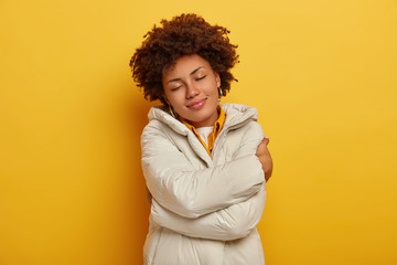 Obraz na płótnie Canvas Beautiful pleased woman enjoys comfort in new winter jacket, embraces herself, keeps eyes shut, feels warm and satisfied, har curly hairstyle, isolated over yellow background. People, clothes concept