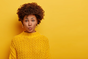 Fototapeta na wymiar Portrait of lovely lady with Afro hairstylre, keeps lips folded, wears silver earrings and knitted yellow sweater has direct gaze at camera stands indoor against bright background copy space for promo
