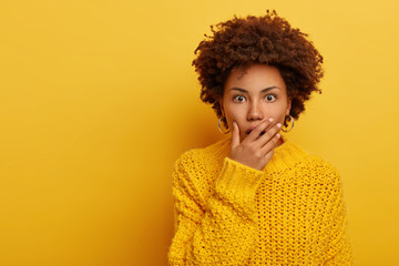 Emotional dark skinned young woman gasps from astonishment, covers opened mouth with palm, looks shocked at camera, wears comfortable knitted yellow sweater in one tone with background. Reaction