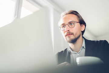 Handsome focused young man sitting by the desk working at the computer. Copy space available on the left and bottom.
