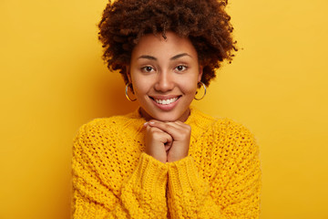 Close up shot of romantic pleasant looking woman keeps both hands under chin, smiles broadly, has nice heartwarming conversation, natural beauty, Afro hairstyle, wears knitted jumper, grins joyfully