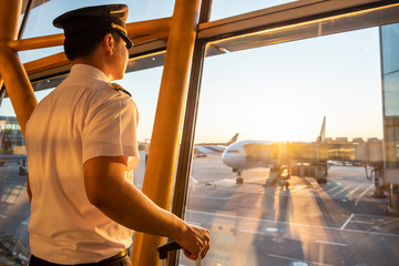 Pilot in uniform standing at boarding gate area in airport terminal, is looking out of window to see ground staff preparing an airplane to be ready for his flight. Seen from back in morning sunrise. © Kawee