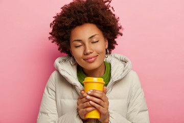 Photo of pleased lovely curly woman wears warm coat with hood, drinks hot coffee during winter time, closes eyes, enjoys good moment, has healthy look, isolated over pink background. People and drink