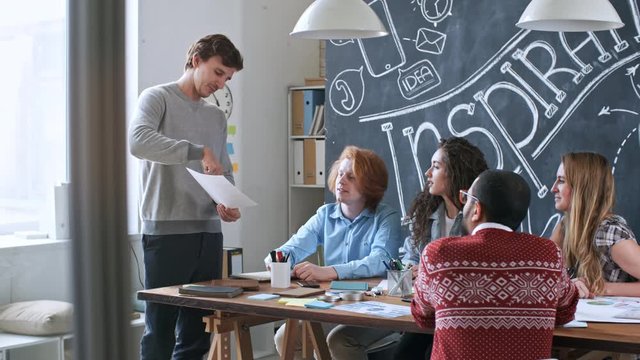 Young mam standing in front of colleagues and presenting business plan to them. Team of creative professionals sitting at meeting table and discussing project.
