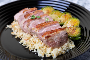 Grilled duck with fried Brussels sprouts, rice from cauliflower on a black plate. Close-up, selective focus.
