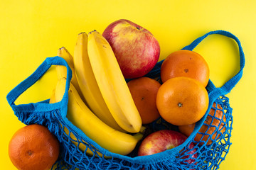 Blue bag string bag with bananas, red apples and tangerines on a bright yellow background. Concept of eco-friendly materials. Close up.