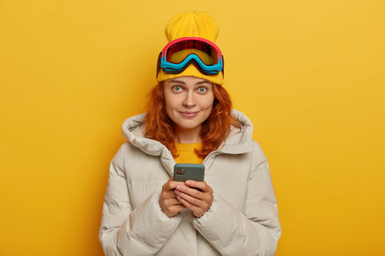 Beautiful ginger woman posts photos in social networks after awesome day, has active rest during winter, holds mobile phone, wears hat, coat and protective ski glasses, poses over yellow wall