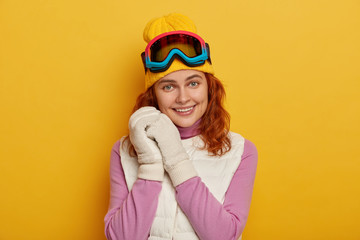 Cheerful ginger woman keeps both hands near face, smiles gently, wears ski goggles, has active rest during winter holidays in resort place, dressed in warm clothes, isolated over yellow studio wall