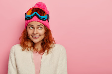 Happy skier girl smiles gently, focused aside, wears pink winter hat with pompon, white soft...