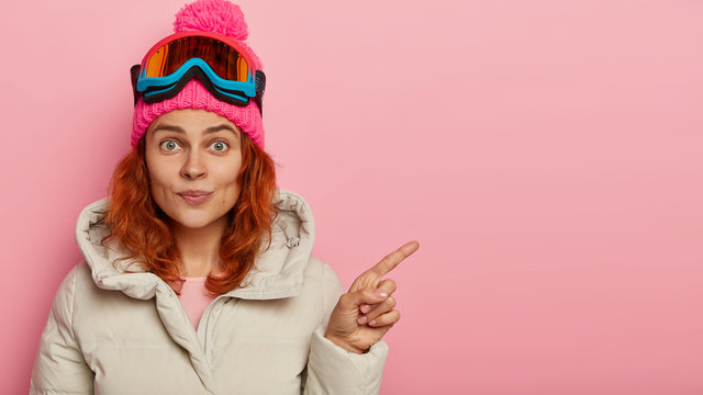 Woman snowboarder dressed in warm clothes, points fore finger on blank space, wears ski glasses, shows something for winter rest, poses against pink background. Extreme sport, leisure, spare time