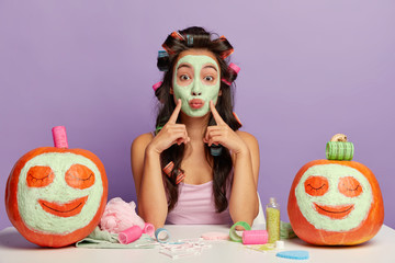 Photo of Asian female model points fore fingers on cheeks, applies purifying face mask, keeps lips...