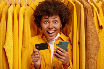 Emotive cheerful shopping woman uses mobile phone application for paying online, holds credit card, happy to get salary, spends all money on clothes, stands between yellow sweaters on hangers © WHstudio Leushin N