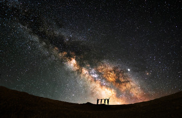 Silhouette of a group of people standing on a hill. Behind them is the beautiful bright Milky Way Galaxy.