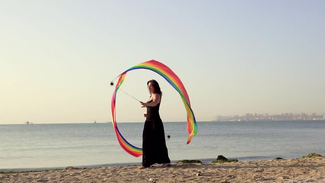 Beautiful young girl juggles poi with the LGBT flag on the beach near sea water at sunset. Sleight of hand and acrobatic with poi. slow motion.