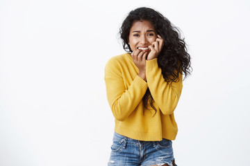 Terrified, scared young african american woman in yellow sweater, biting fingers, frowning and...