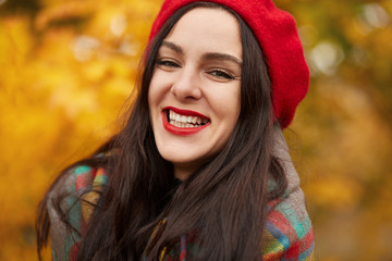 Beautiful elegant woman standing in park in autumn, young female with charming smile wearing red beret and wrapped warm checkered blanket, looking at camera a nd laughing, expresses happyness.