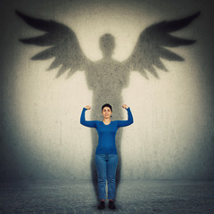 Casual confident woman flexing arm muscles as casting a hero shadow with angel wings on a dark room wall. Super power, inner strength ambition and leadership. Female independence and energy concept.