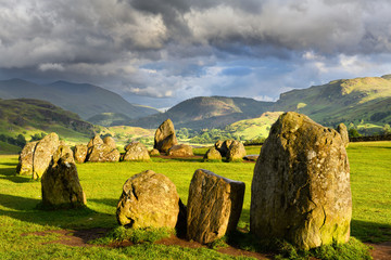 Sun on Castlerigg Stone Circle under dark clouds on summer solstice eve with Naddle village in Cumbrian Mountains Lake District Keswick England