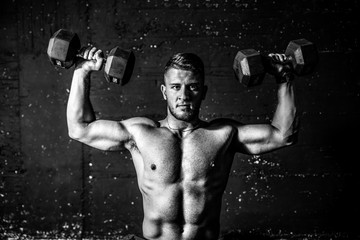 Young fit strong sweaty man shoulders workout training with two dumbbells in the gym dark image...