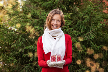 Attractive happy smiling young woman with Christmas gift box on her hands in knitted mittens and scarf on fir tree background with golden lightning bokeh. Celebration New Year concept.