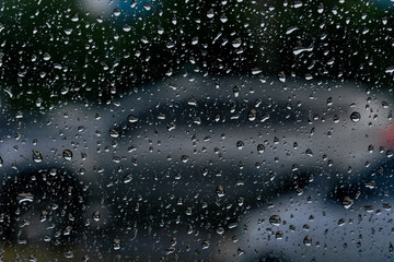 A glass with rain drops that have a backdrop as a car in a parking lot.