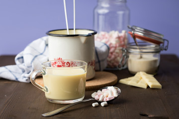 Zubereitung von heißer weiße Schokolade im Milchtopf mit Kakao Pulver und Marshmallows in Küche auf Holz Arbeitsfläche dunkel und Lila Hintergrund