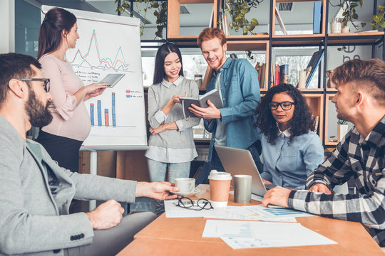 Startupers working together at office sitting looking for new ideas on laptop while pregnant woman browsing digital tablet and other colleagues reading books cheerful
