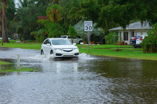 Heavy flooding and storm surge in residential neighborhood with a car driving through deep splashing water in the flooded street in front of houses with Speed Limit sign on side of the road.