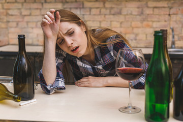 Young sad and wasted alcoholic woman sitting at kitchen couch drinking red wine, completely drunk...