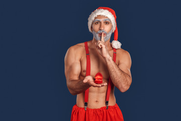 Christmas Freestyle. Young Santa Claus bare muscular upper body in hat standing isolated on blue holding proposal ring showing silence gesture surprise