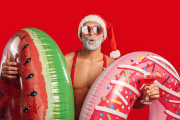 Christmas Freestyle. Young Santa Claus bare muscular upper body in hat and sunglasses standing isolated on red with swim rings and cocktail surprised