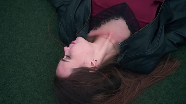 A beautiful and passionate girl in a biker jacket lies on the floor of a basketball or tennis court. A young woman is lying on the green surface of a sports court. Close-up.