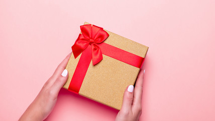Woman manicured hands holding red and golden wrapped present or gift box on pastel pink background, copy space, top view, flat lay. Background for Valentine's Day, Mother's Day.
