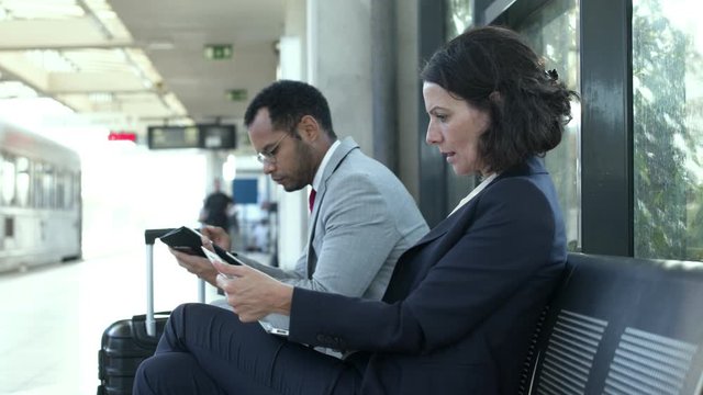 Focused business people with digital devices. Multiethnic businessman and businesswoman using laptop and digital tablet while sitting and waiting at railway station. Digital devices concept