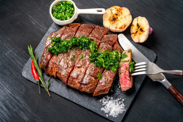 Ready to eat black Angus beef rib eye steak sliced with herbs, garlic and sauce on slate Board. Ready meal for dinner on black wooden background
