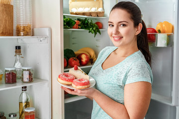 Body Care. Chubby girl standing in kitchen near fridge holding plate with doughnuts and cheesecake smiling friendly to camera