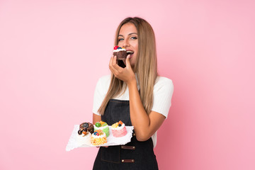 Young blonde woman over isolated pink background holding mini cakes and eating it