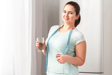 Body Care. Chubby girl standing in kitchen near window with tape measure around neck holding glass of water smiling joyful