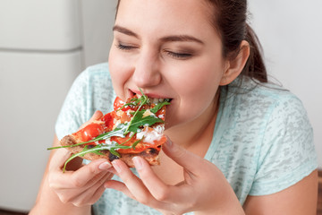 Binge Eating. Chubby girl sitting at kitchen table eating pizza closed eyes delightful close-up