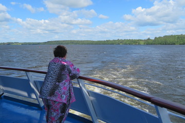 European river cruise, a big female passenger look the landscape from the stern desk of the ship on water and blue sky with clouds background on Sunny summer day