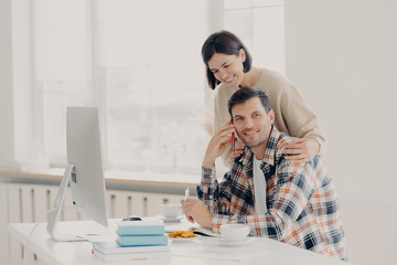 Obraz premium Caring woman helps her husband entrepreneur who has telephone conversation, tries to soleve working issues, writes information in notepad and works on computer. Family couple manage finances