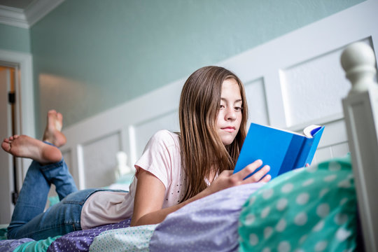 Adolescent teen girl reading a book while lying in bed at home in her bedroom. Lifestyle photo